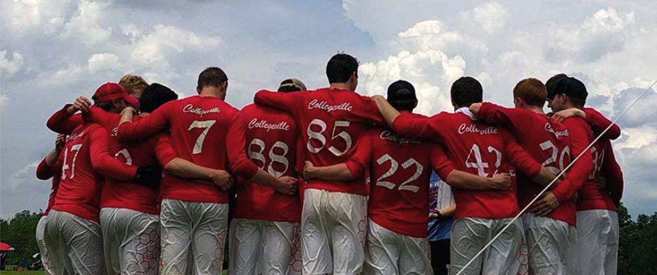 A sports team huddles with their arms around each other's shoulders. They are all wearing red jerseys with white numbers and lettering that say "Collegeville" on the back. The sky is cloudy, and the scene suggests a sense of unity and teamwork.